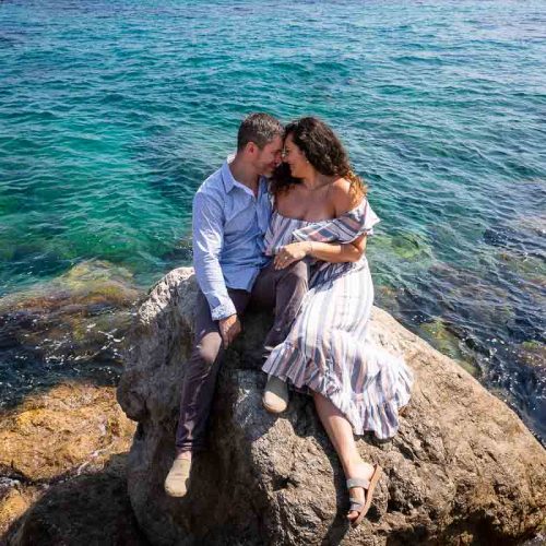 Capri engagement photo session. Couple sitting down on the rocks surrounded by crystalline emerald green blue water all around