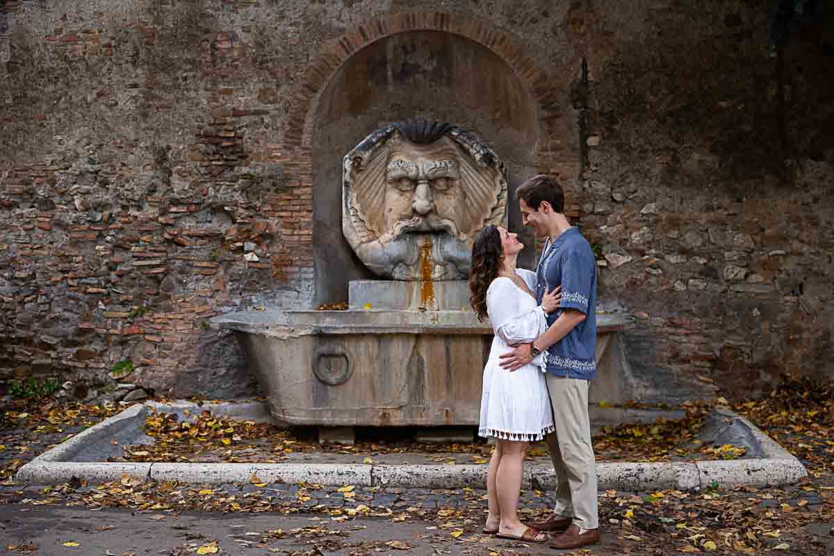 Full figure picture frame of a couple posing in front of the ancient roman water fountain found in front of the entrance to the Giardino degli Aranci park 