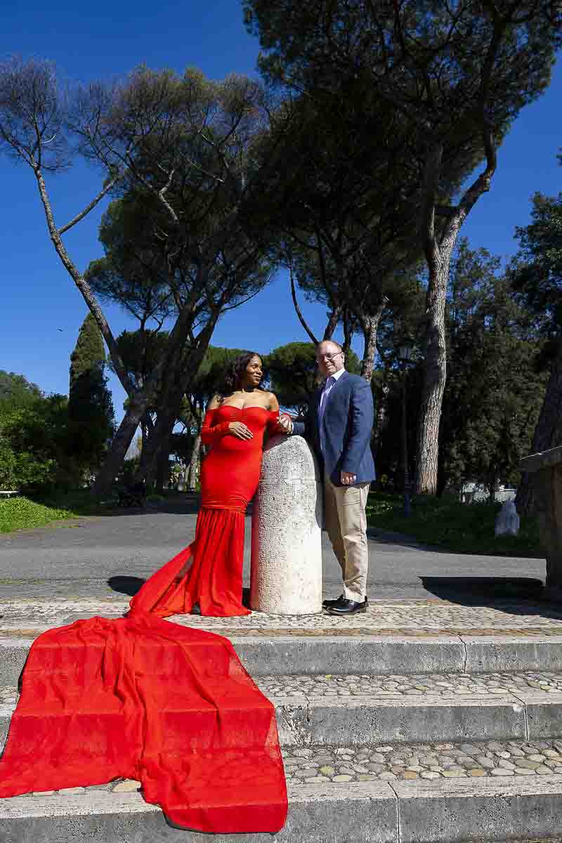 Posed couple with a long red dress tail falling over white marble steps in a park with Mediterranean pine trees photographer above by a Rome lifestyle photographer 